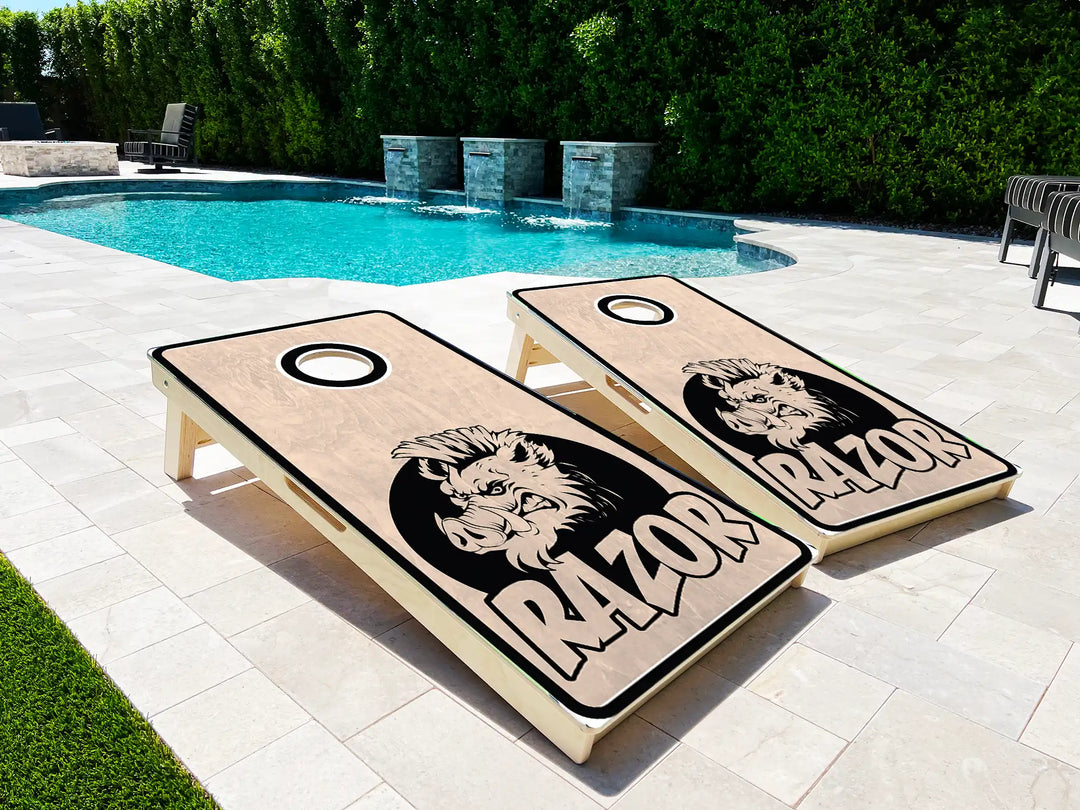 Two cornhole boards with a boar design and 'RAZOR' text on a patio near a pool.