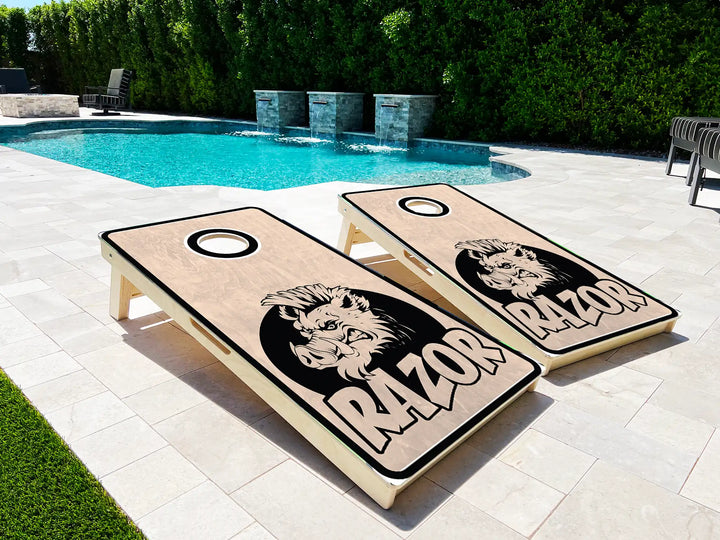 Two cornhole boards with a boar design and 'RAZOR' text on a patio near a pool.