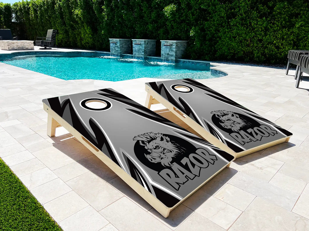 Two cornhole boards with a lion logo and 'Razor' text on a patio near a pool.