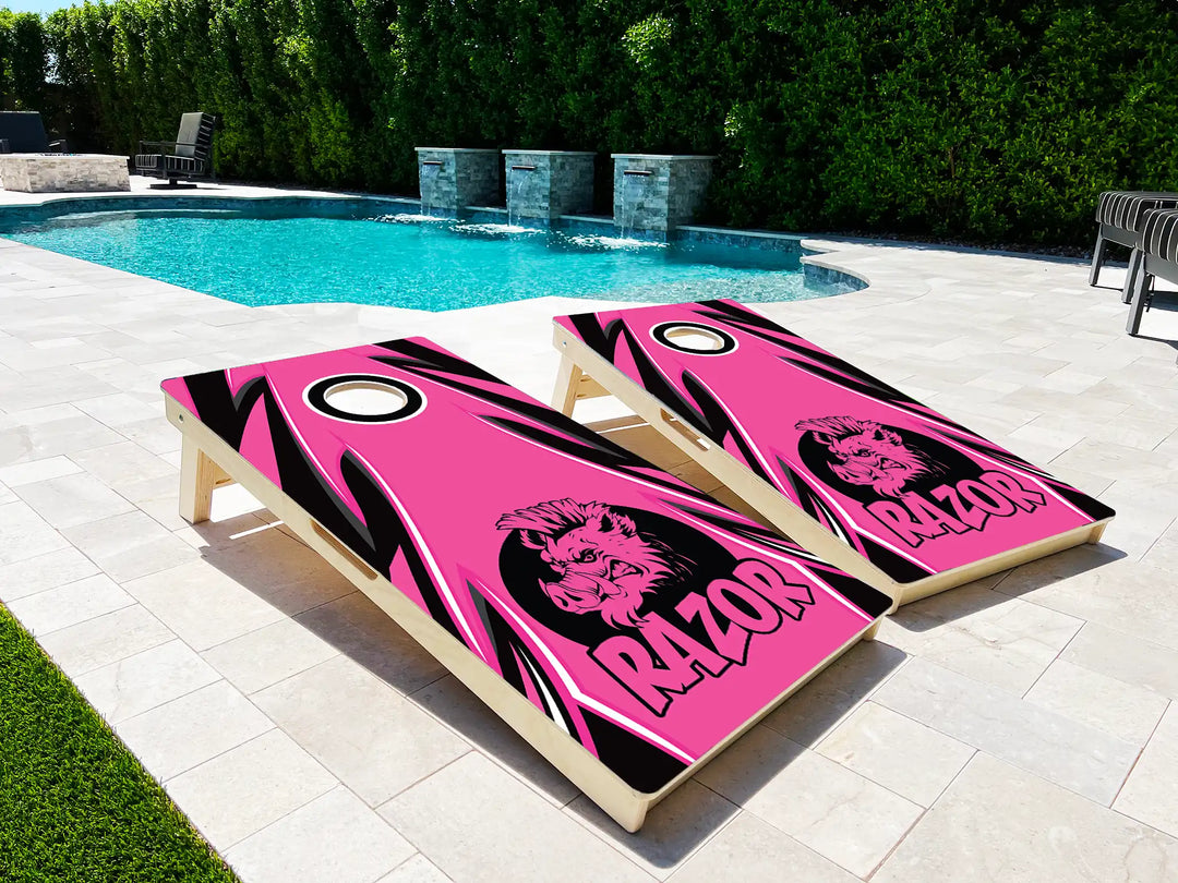 Two pink and black cornhole boards with a logo and 'Razor' text on a patio near a pool.