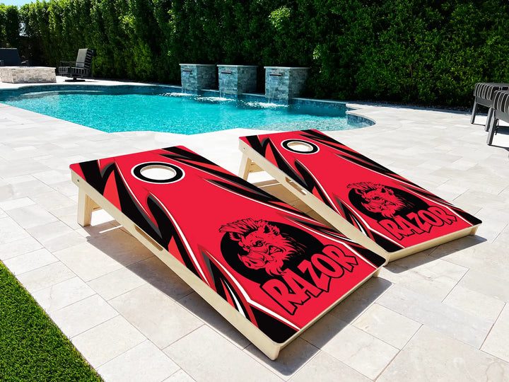 Two cornhole boards with a 'Razor' design on a patio near a pool.